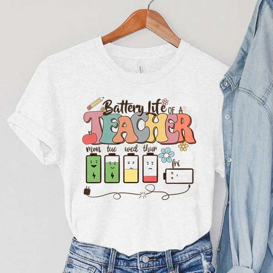 Ivory 'Battery Life of a Teacher' t-shirt hanging on a wooden hanger. The shirt showcases playful graphics that humorously illustrate a teacher's energy levels throughout the day, making it a cute and trendy choice for educators. Brighten your wardrobe and shop today at teachingteez.com!