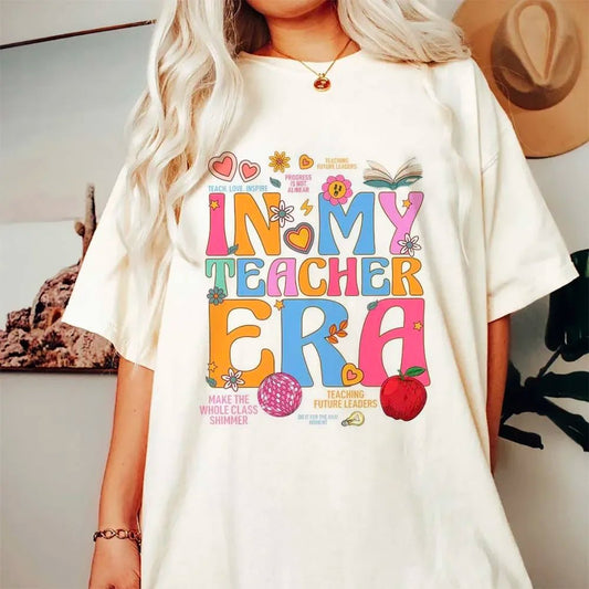 White t-shirt with a colorful "In My Teacher Era" design featuring retro-style letters, hearts, flowers, an apple, and a glittery disco ball. The graphic also includes fun phrases like "Make the Whole Class Shimmer" and "Teaching Future Leaders." Perfect for teachers who love a vibrant, playful look. Available at TeachingTeez.com.