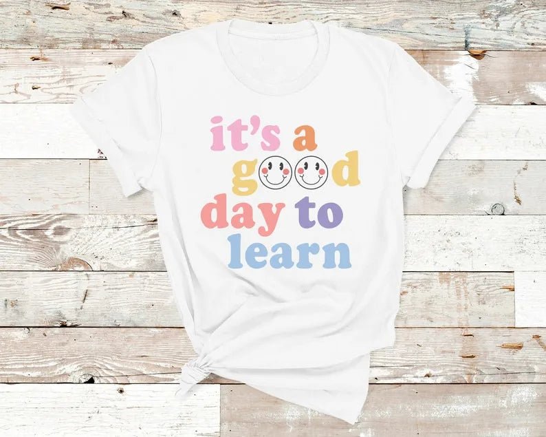 White shirt Flat lay of the "It's a Good Day to Learn" t-shirt featuring colorful, playful lettering with smiley faces in the "O"s of "Good," crafted from soft fabric for comfort—shop today and spread positivity!