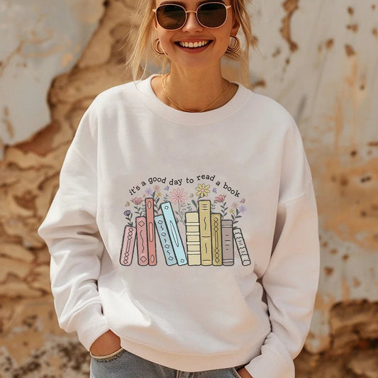 Woman wearing a white sweatshirt with a charming "It's a Good Day to Read a Book" design, perfect for book lovers and teachers. The graphic features pastel-colored books with flowers blooming from their pages, adding a whimsical touch to this cozy teacher sweatshirt. Ideal for showing off a love of reading. Available at TeachingTeez.com.