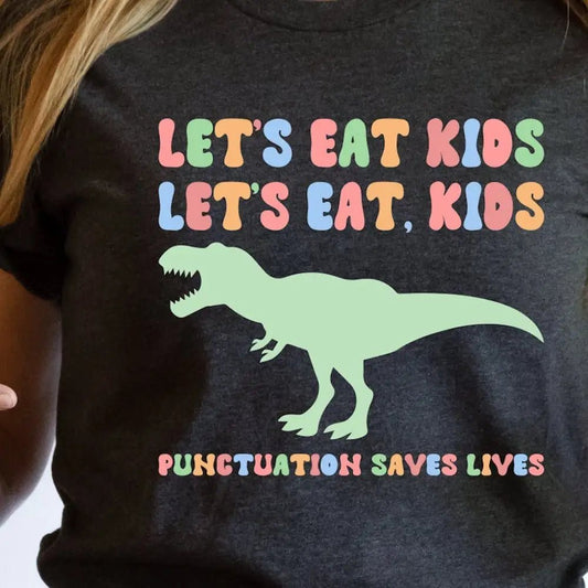 Charcoal gray t-shirt featuring the humorous phrase "Let’s Eat Kids, Let’s Eat, Kids" in colorful, retro-style letters, along with a playful green T-Rex graphic. The tagline "Punctuation Saves Lives" adds a clever grammar twist, making it a fun choice for English teachers and grammar enthusiasts. This cute teacher t-shirt is perfect for sparking conversation about punctuation in the classroom. Available at TeachingTeez.com.