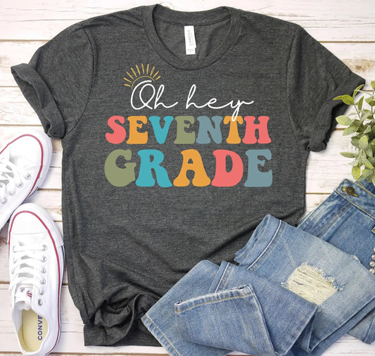 Flat lay of the grey "Oh Hey Seventh Grade" t-shirt from TeachingTeez.com, featuring the playful phrase "Oh Hey Seventh Grade" in colorful retro fonts and a cheerful sun illustration. This cute teacher t-shirt showcases a fun and vibrant design, perfect for teachers looking to brighten their wardrobe with stylish teacher shirts. Shop now for trendy teacher tshirts that celebrate seventh-grade enthusiasm!