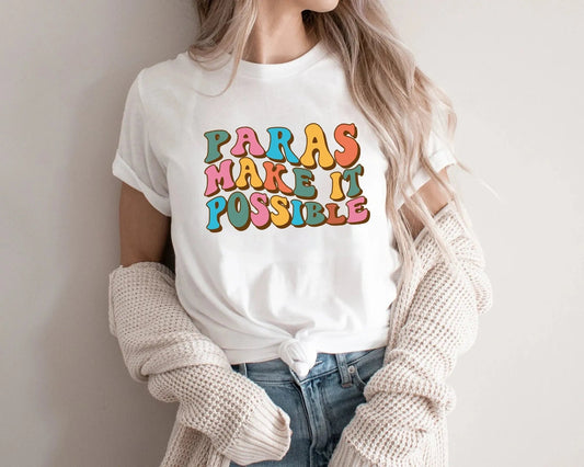 White t-shirt with the phrase "Paras Make It Possible" in colorful, retro-style bubble letters. The shirt celebrates paraprofessionals with a fun and vibrant design, perfect for showing appreciation for paras in schools. Styled with jeans and a cozy knit cardigan, this cute paraprofessional t-shirt is ideal for casual wear. Available at TeachingTeez.com.