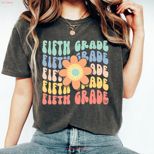 Charcoal gray t-shirt with a colorful, retro "Fifth Grade" design. The words "Fifth Grade" are stacked vertically in bright colors, each line using a fun, nostalgic font. A large retro flower is centered on the design, adding a playful, vintage flair—perfect for fifth-grade teachers who want to showcase their grade-level pride with style. Available at TeachingTeez.com.