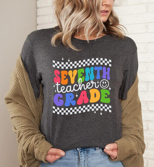 A woman wearing a dark gray t-shirt with a colorful retro design that reads "Seventh Grade Teacher" in bold, playful letters. The shirt features a checkered pattern at the top and bottom, adding a vintage vibe, with each letter in a different bright color—red, orange, green, blue, and purple. Small star accents and a smiley face bring a fun, cheerful touch to the design, making it a stylish and cute teacher t-shirt ideal for seventh-grade educators. She is casually styled with an open cardigan and jeans.