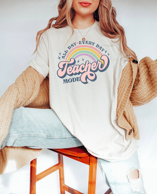 A woman wearing a beige "All Day Every Day Teacher Mode" T-Shirt from TeachingTeez.com, featuring a retro pastel rainbow and bold, playful text. This trendy teacher shirt is perfect for educators showing off their love for teaching with style. Get this comfortable, inspirational t-shirt from TeachingTeez.com and stay in teacher mode all day!